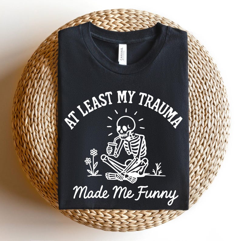 K&ouml;nnte beinhalten: Schwarzes T-Shirt mit dem Text "AT LEAST MY TRAUMA Made Me Funny" und einer wei&szlig;en Grafik eines sitzenden Skeletts, das aus einer Tasse trinkt. Das T-Shirt liegt auf einer runden, gewebten Oberfl&auml;che.