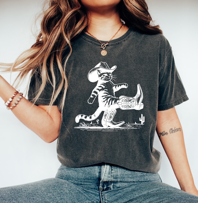 May include: Dark gray t-shirt featuring a white illustration of a cat wearing a cowboy hat and boots. The cat is walking on a line with cacti. The shirt is a comfortable, casual style.