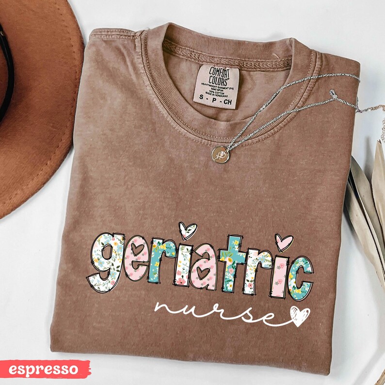 Pode incluir: Camiseta marrom com as palavras "geriatric nurse" em um design floral. A camisa tem gola redonda e mangas curtas. Um colar de prata com um pequeno pingente est&aacute; drapeado sobre a camisa.