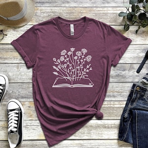 K&ouml;nnte beinhalten: Ein maroonfarbenes T-Shirt mit einer wei&szlig;en Grafik eines Buches, aus dem Blumen wachsen.