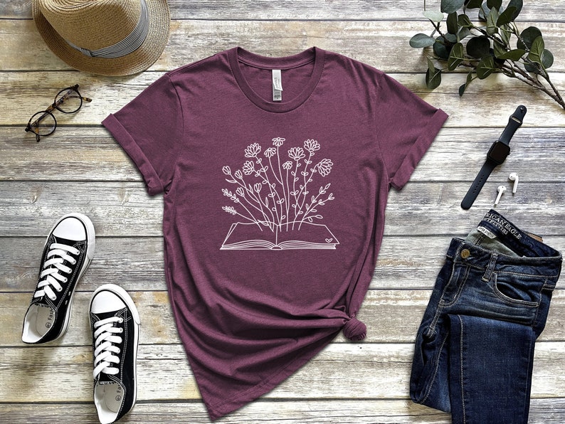 K&ouml;nnte beinhalten: Ein maroonfarbenes T-Shirt mit einer wei&szlig;en Grafik eines Buches, aus dem Blumen wachsen.