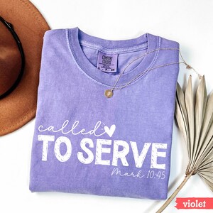 Pu&ograve; includere: Una maglietta Comfort Colors viola con le parole "called TO SERVE" in corsivo bianco, un piccolo cuore e il testo "Mark 10:45". Una collana d'oro e un cappello marrone sono visibili. Una foglia di palma secca &egrave; a destra.