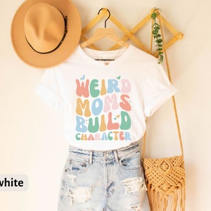 May include: White t-shirt with the text "Weird Moms Build Character" in colourful, wavy letters. The shirt is hanging on a wooden rack with a straw hat and a woven bag.