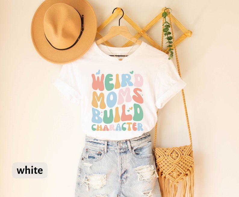 May include: White t-shirt with the text "Weird Moms Build Character" in colourful, wavy letters. The shirt is hanging on a wooden rack with a straw hat and a woven bag.