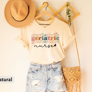 Może przedstawiać: Naturalny kolorowy t-shirt z napisem "Geriatric Nurse" w kolorowych, ułożonych na sobie literach. Koszulka wisi na drewnianym wieszaku z plecioną torbą i kapeluszem.