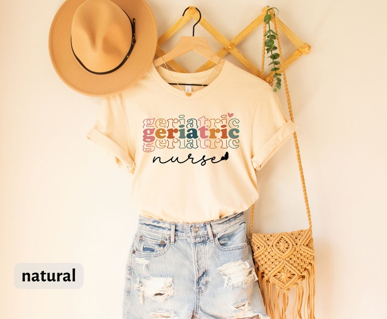 Może przedstawiać: Naturalny kolorowy t-shirt z napisem "Geriatric Nurse" w kolorowych, ułożonych na sobie literach. Koszulka wisi na drewnianym wieszaku z plecioną torbą i kapeluszem.
