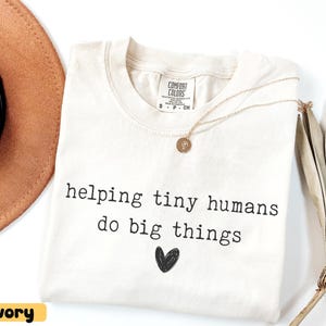 May include: Ivory-colored t-shirt with the text "helping tiny humans do big things" and a black heart graphic. A gold necklace with a pendant rests on the shirt. A brown hat is visible in the background.