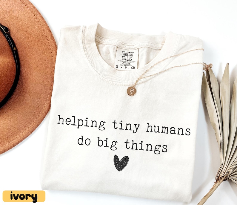 May include: Ivory-colored t-shirt with the text "helping tiny humans do big things" and a black heart graphic. A gold necklace with a pendant rests on the shirt. A brown hat is visible in the background.