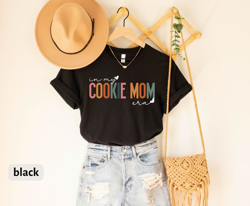 May include: Black t-shirt with the text "in my cookie mom era" in rainbow colors. The shirt is hanging on a wooden rack with a straw bag and a plant.