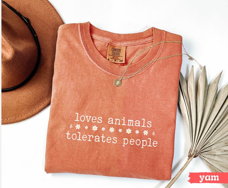 May include: A coral-colored t-shirt with the text "loves animals tolerates people" in white lettering. The text is surrounded by small white flower graphics.