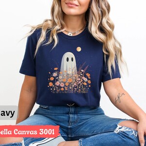 Pu&ograve; includere: T-shirt blu navy con un fantasioso disegno di un fantasma in mezzo a un campo di fiori sotto la luna piena. Il fantasma ha due occhi ovali neri. Il testo "navy" e "Bella Canvas 3001" sono visibili nell'immagine.