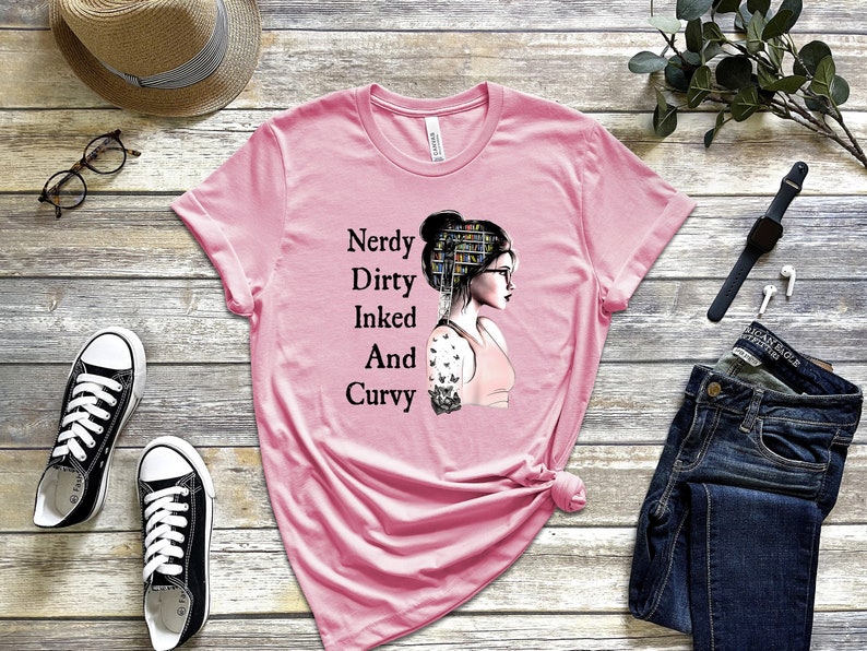 Op de afbeelding: Roze T-shirt met een afbeelding van een vrouw met boeken op haar hoofd en een tatoeage op haar arm. De tekst op het shirt luidt "Nerdy Dirty Inked And Curvy".
