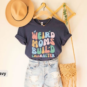May include: Navy blue t-shirt featuring the text "Weird Moms Build Character" in colourful, retro-style lettering. The shirt is displayed on a mannequin wearing denim shorts and a straw bag.