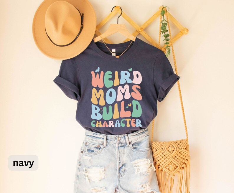 May include: Navy blue t-shirt featuring the text "Weird Moms Build Character" in colourful, retro-style lettering. The shirt is displayed on a mannequin wearing denim shorts and a straw bag.