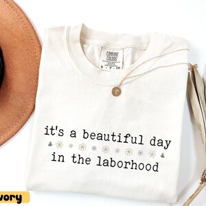 May include: Ivory-colored Comfort Colors t-shirt with the phrase "it's a beautiful day in the laborhood" in black font. The shirt features small flower and butterfly graphics. A gold necklace with a pendant rests on the shirt.