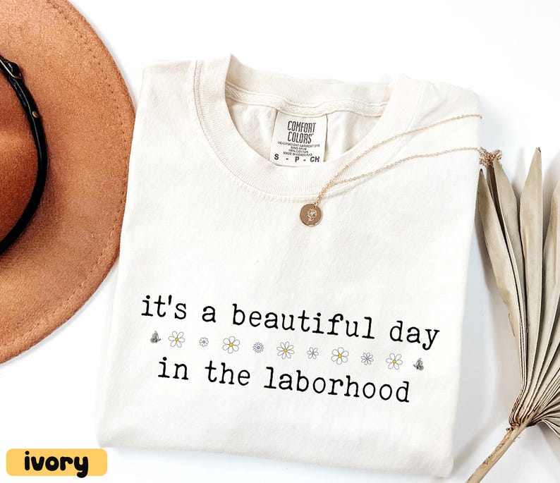 May include: Ivory-colored Comfort Colors t-shirt with the phrase "it's a beautiful day in the laborhood" in black font. The shirt features small flower and butterfly graphics. A gold necklace with a pendant rests on the shirt.