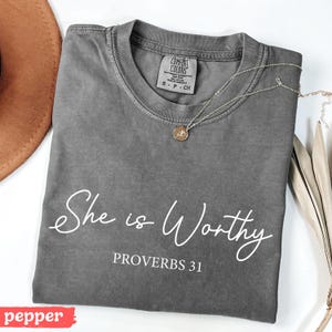 May include: A gray Comfort Colors t-shirt with the text "She is Worthy" and "PROVERBS 31" in white script. A gold necklace with a pendant is draped across the shirt. A brown hat and dried leaves are in the background.