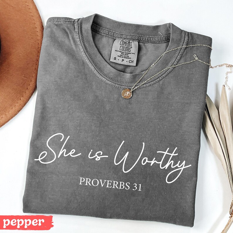 May include: A gray Comfort Colors t-shirt with the text "She is Worthy" and "PROVERBS 31" in white script. A gold necklace with a pendant is draped across the shirt. A brown hat and dried leaves are in the background.