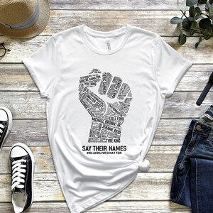 Puede incluir: Camiseta blanca con un gr&aacute;fico de pu&ntilde;o negro hecho de nombres. El pu&ntilde;o est&aacute; levantado en el aire. El texto debajo del pu&ntilde;o dice "Say Their Names #BlackLivesMatter".