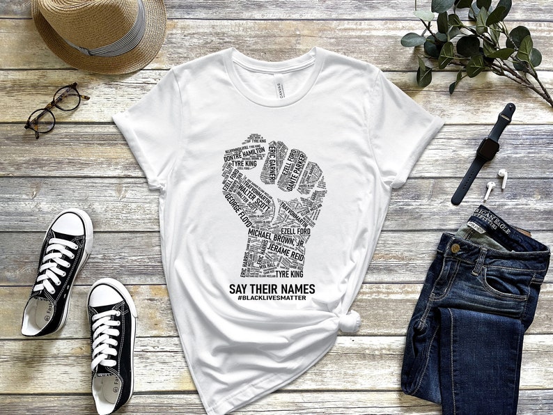Puede incluir: Camiseta blanca con un gr&aacute;fico de pu&ntilde;o negro hecho de nombres. El pu&ntilde;o est&aacute; levantado en el aire. El texto debajo del pu&ntilde;o dice "Say Their Names #BlackLivesMatter".