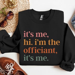 May include: Black sweatshirt with the text "it's me, hi. i'm the officiant, it's me." in colourful letters.