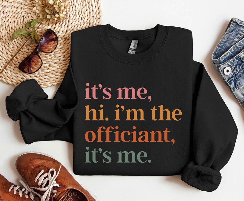 May include: Black sweatshirt with the text "it's me, hi. i'm the officiant, it's me." in colourful letters.