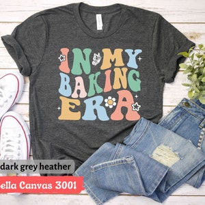 May include: A dark grey heather Bella Canvas 3001 t-shirt with the text "In My Baking Era" in colorful retro-style lettering. The text is decorated with a butterfly, a flower, and a star.