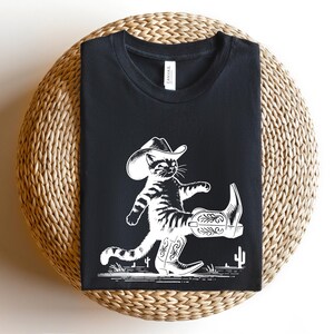 May include: Black t-shirt featuring a white illustration of a cat wearing a cowboy hat and boots. The cat is mid-stride, with one leg raised, and a cactus is in the background. The shirt is folded on a woven, circular surface.