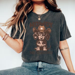 Puede incluir: Camiseta gris oscuro con un perro salchicha marr&oacute;n que lleva un sombrero de vaquero con una estrella blanca. El texto "HOWDY DACHSHUND" est&aacute; impreso encima y debajo de la ilustraci&oacute;n del perro. La camiseta tiene un aspecto vintage.