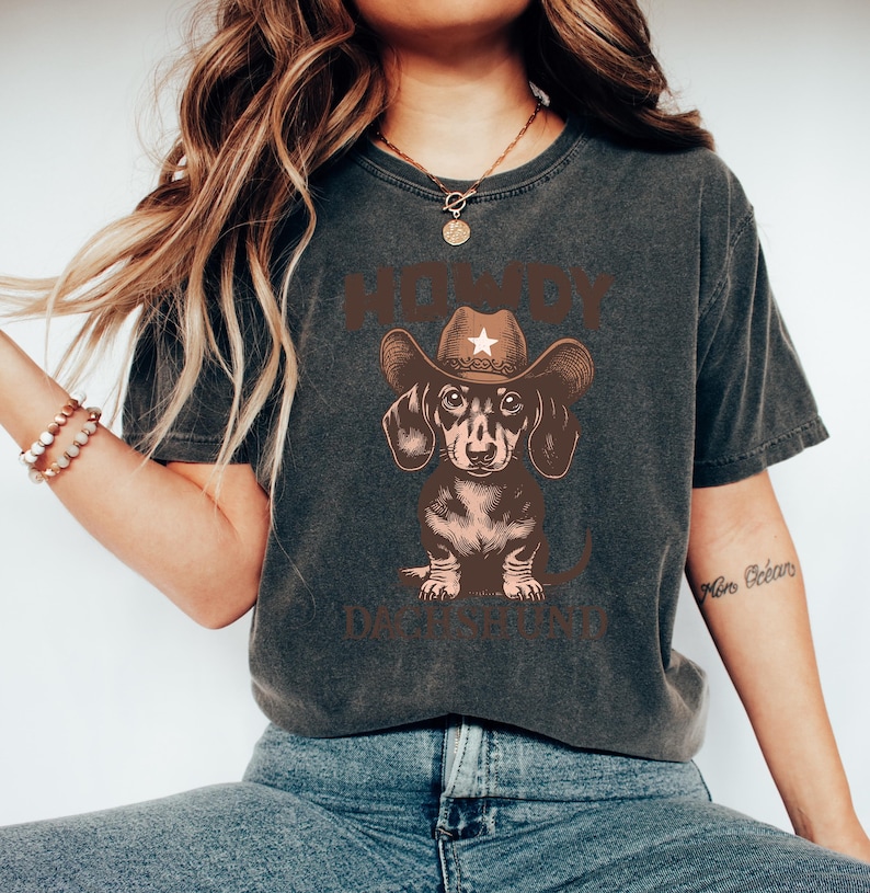 Puede incluir: Camiseta gris oscuro con un perro salchicha marr&oacute;n que lleva un sombrero de vaquero con una estrella blanca. El texto "HOWDY DACHSHUND" est&aacute; impreso encima y debajo de la ilustraci&oacute;n del perro. La camiseta tiene un aspecto vintage.