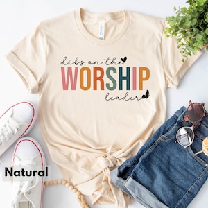 Puede incluir: Una camiseta beige con el texto "dibs on the WORSHIP leader" en letras de colores. La camiseta est&aacute; doblada y colocada sobre una superficie blanca con un par de zapatillas blancas, unos pantalones cortos de mezclilla azul y un collar de cuentas de madera.