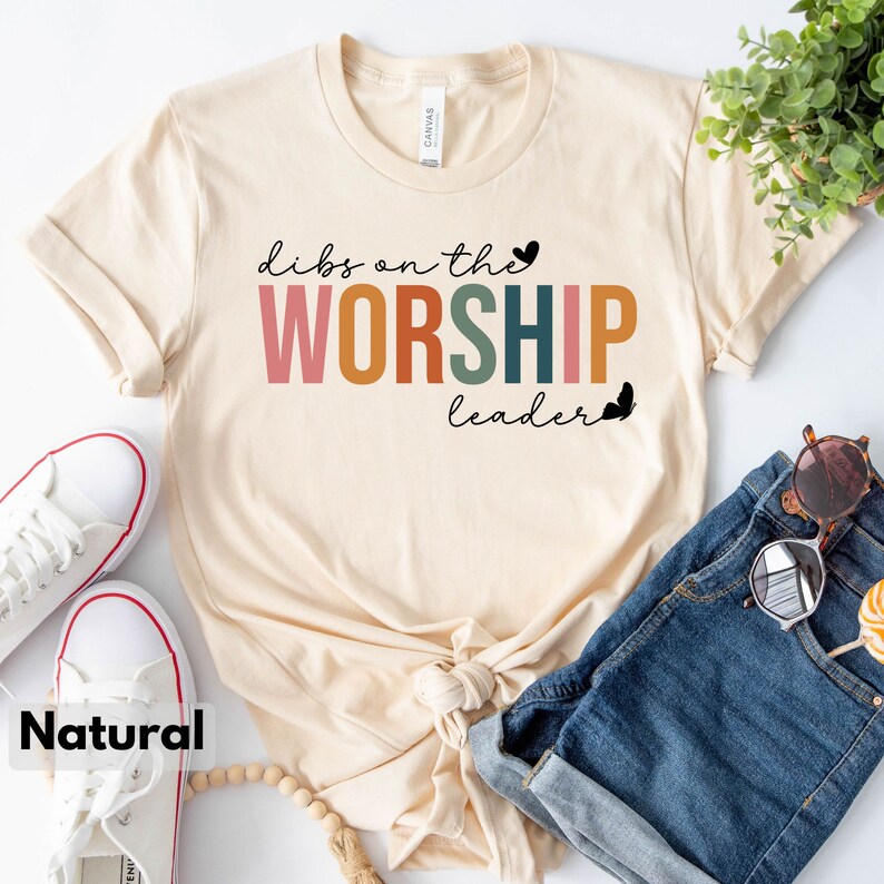 Puede incluir: Una camiseta beige con el texto "dibs on the WORSHIP leader" en letras de colores. La camiseta est&aacute; doblada y colocada sobre una superficie blanca con un par de zapatillas blancas, unos pantalones cortos de mezclilla azul y un collar de cuentas de madera.