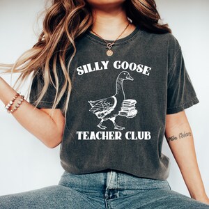 May include: A dark gray t-shirt with a white graphic of a goose carrying books and the text "Silly Goose Teacher Club".