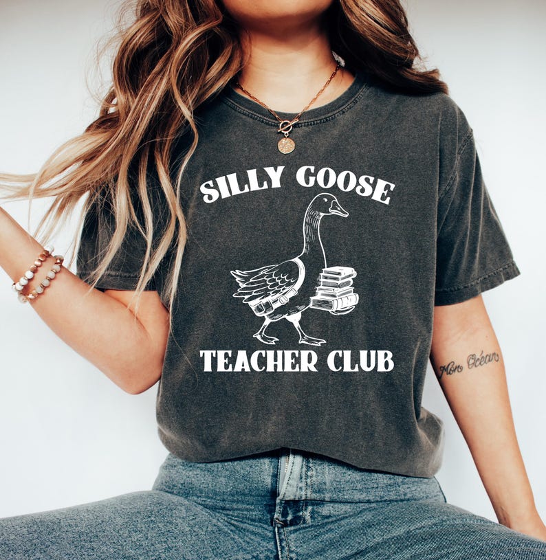 May include: A dark gray t-shirt with a white graphic of a goose carrying books and the text "Silly Goose Teacher Club".
