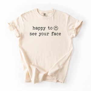 May include: A cream colored t-shirt with the text "happy to see your face" and a smiley face graphic printed on the front.