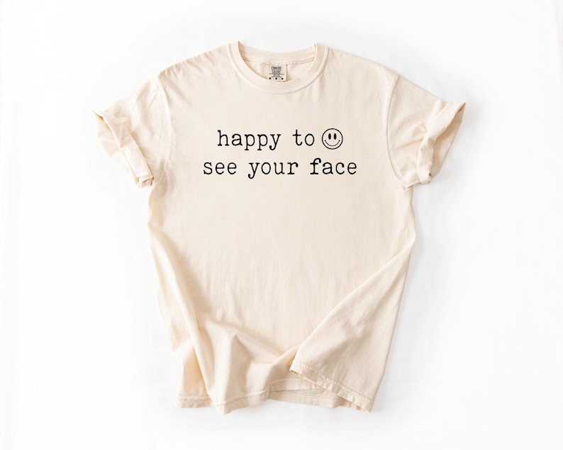May include: A cream colored t-shirt with the text "happy to see your face" and a smiley face graphic printed on the front.