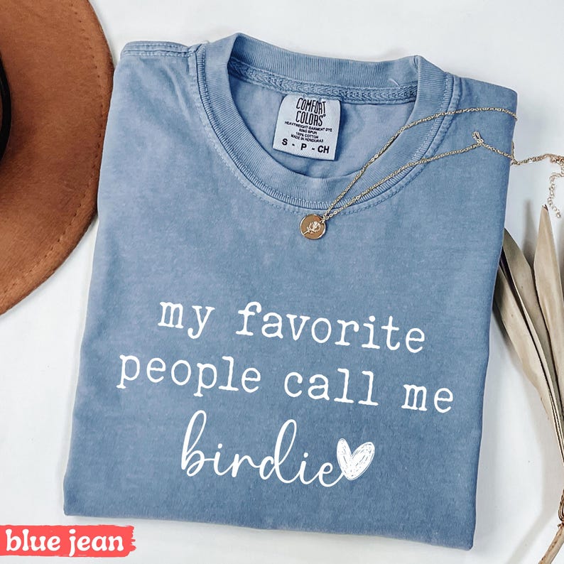 May include: Blue jean coloured t-shirt with the text "my favourite people call me birdie" in white script. A gold necklace with a pendant is draped across the shirt. The shirt is folded and has a tag that says "Comfort Colors".