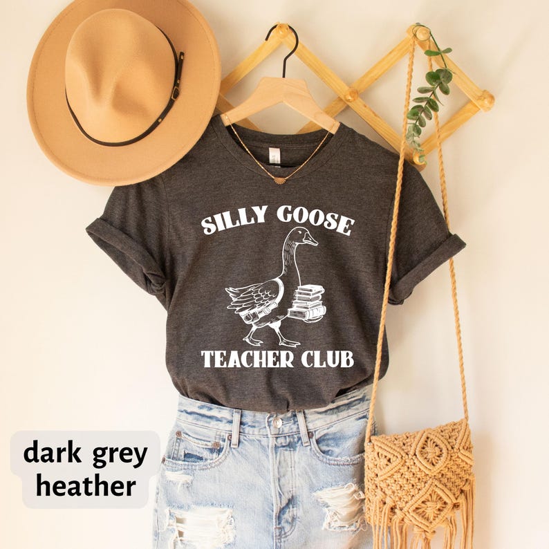 May include: Dark grey heather V-neck t-shirt with a white graphic of a goose carrying books. The text "Silly Goose Teacher Club" is printed on the shirt.