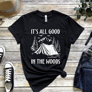 May include: Black t-shirt with white text that reads "It's all good in the woods". The design features a tent, trees, and mountains.