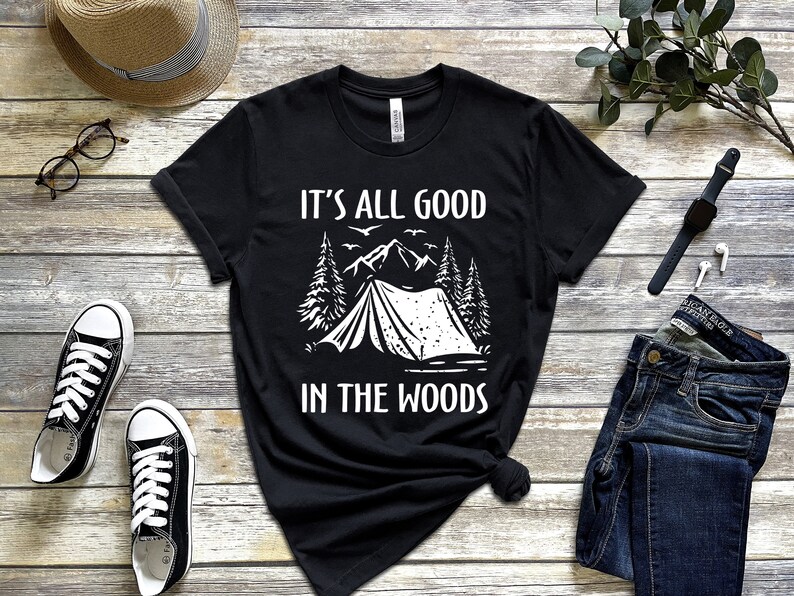 May include: Black t-shirt with white text that reads "It's all good in the woods". The design features a tent, trees, and mountains.