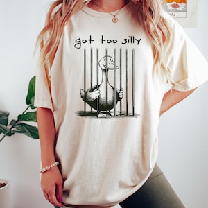 May include: A white t-shirt with a black and white illustration of a duck in a cage. The text "got too silly" is printed above the illustration.