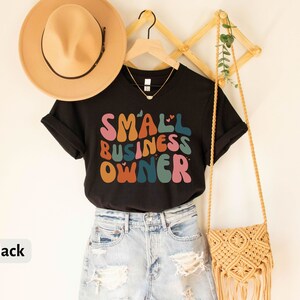 May include: Black t-shirt with the text "SMALL BUSINESS OWNER" in colorful, retro-style lettering. The shirt is being modeled with a straw hat and a crocheted bag.