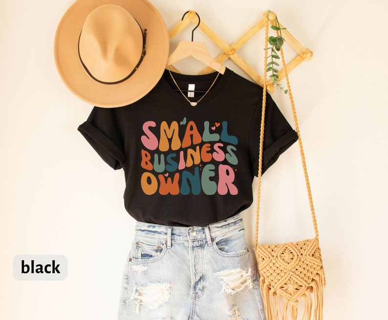 May include: Black t-shirt with the text "SMALL BUSINESS OWNER" in colorful, retro-style lettering. The shirt is being modeled with a straw hat and a crocheted bag.