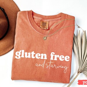 Puede incluir: Una camiseta color &ntilde;ame con las palabras "gluten free and starving" en blanco. La camiseta est&aacute; adornada con un collar dorado, un sombrero marr&oacute;n y una hoja de palma seca. La camiseta tiene una etiqueta Comfort Colors.