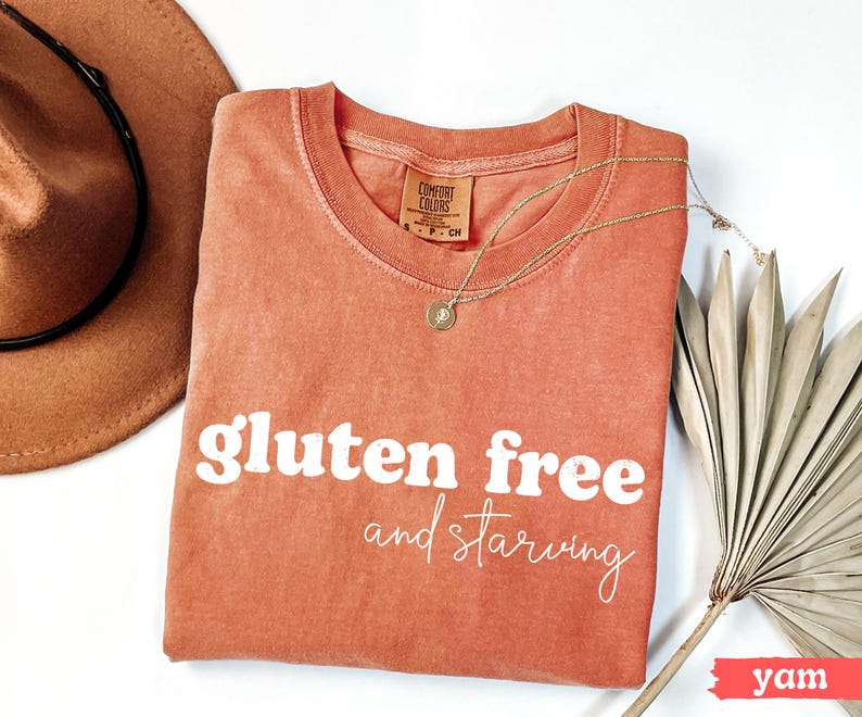 Puede incluir: Una camiseta color &ntilde;ame con las palabras "gluten free and starving" en blanco. La camiseta est&aacute; adornada con un collar dorado, un sombrero marr&oacute;n y una hoja de palma seca. La camiseta tiene una etiqueta Comfort Colors.