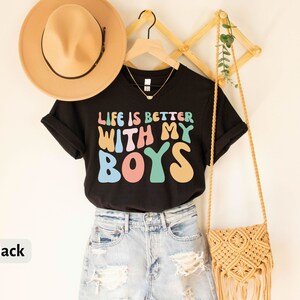 K&ouml;nnte beinhalten: Schwarzes T-Shirt mit dem Text "Life is better with my boys" in Regenbogenfarben. Das Shirt h&auml;ngt an einem Holzgestell mit einer Strohtasche und einem Hut.