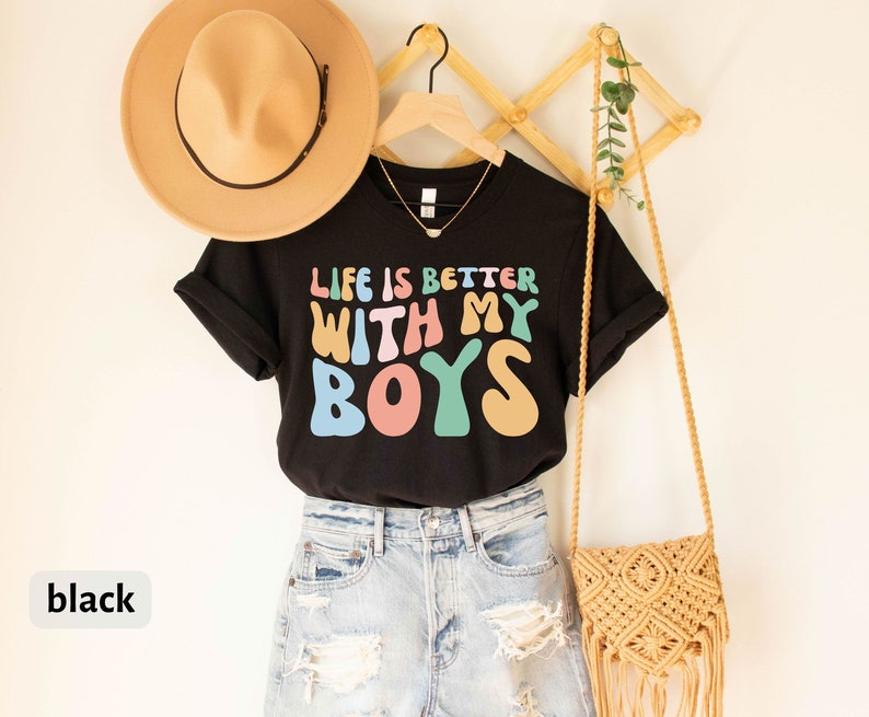 K&ouml;nnte beinhalten: Schwarzes T-Shirt mit dem Text "Life is better with my boys" in Regenbogenfarben. Das Shirt h&auml;ngt an einem Holzgestell mit einer Strohtasche und einem Hut.