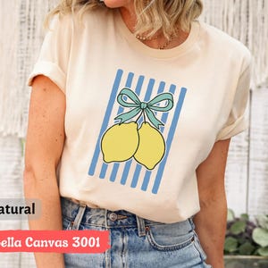 May include: A cream colored t-shirt with a blue and white striped design featuring two yellow lemons with a blue bow. The text "Bella Canvas 3001" and "natural" are printed on the bottom of the shirt.
