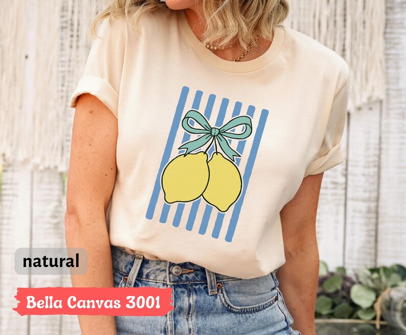 May include: A cream colored t-shirt with a blue and white striped design featuring two yellow lemons with a blue bow. The text "Bella Canvas 3001" and "natural" are printed on the bottom of the shirt.