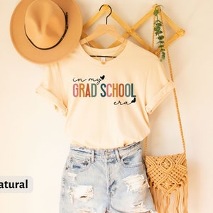 May include: A beige t-shirt with the text "in my grad school era" in rainbow colors and black butterflies. The shirt is hanging on a wooden hanger with a straw bag and a brown hat.